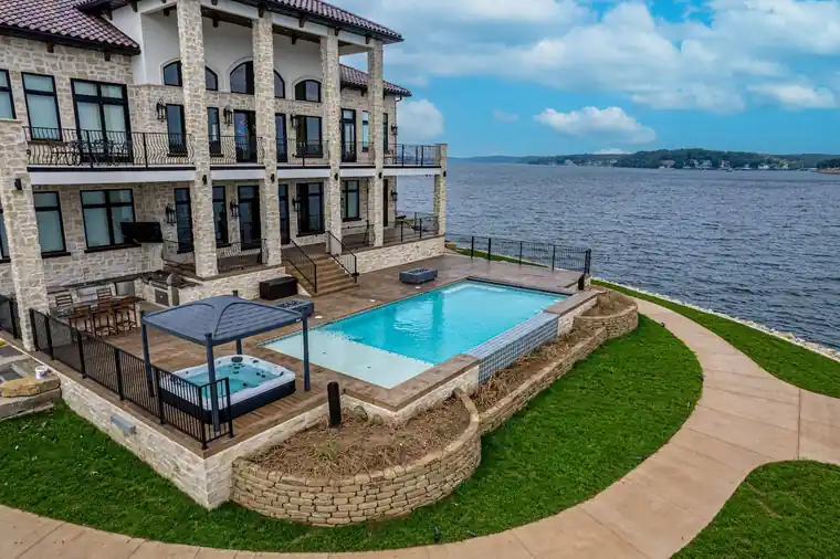 Lakeside stone home at the Lake of the Ozarks with an elevated rectangular pool, a hot tub under a gazebo and the lake beyond