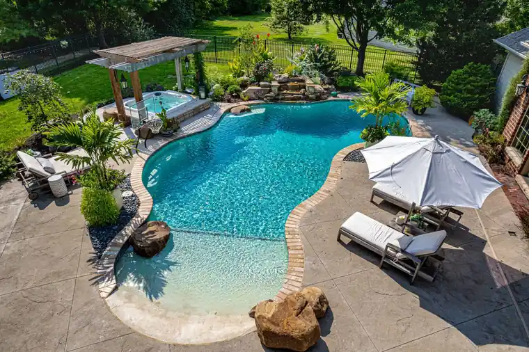 Aerial view of a freeform pool with a beach entry, rock waterfall, raised spa under a pergola, brick coping and a stamped concrete deck